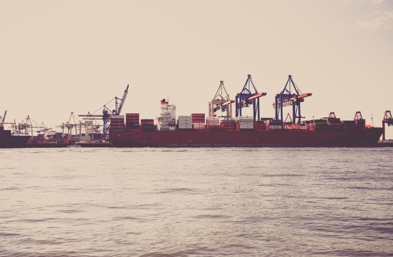services-04 A cargo ship loaded with containers docked at a seaport under clear skies.